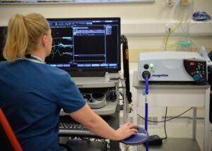 A healthcare professional in scrubs interacts with a computer displaying medical data while using a device labeled 'magstim' in a clinical setting.