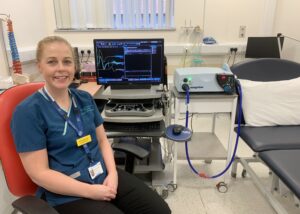 A healthcare professional sitting in a clinical setting, smiling in front of a computer monitor displaying data, with a medical device on a nearby table.