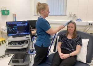 A healthcare professional in a blue uniform is using a device to conduct an examination on a seated woman in a black and white patterned shirt. A medical monitor displaying data is visible in the background.